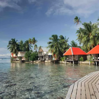 Tropical bungalows over water with palm trees and a wooden walkway.