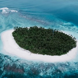 Aerial view of a small tropical island with dense palm trees, surrounded by turquoise water and white sandy beach.