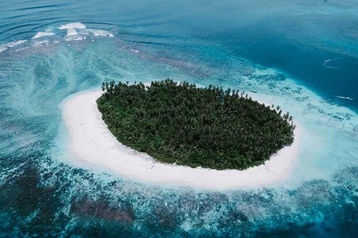 Aerial view of a small tropical island with dense palm trees, surrounded by turquoise water and white sandy beach.