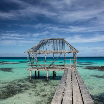 Wooden dock leading to a dilapidated hut over clear turquoise water.