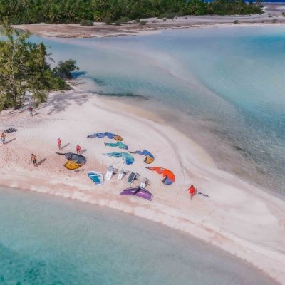 Aerial view of a sandy beach with colorful kites and trees near clear blue water.