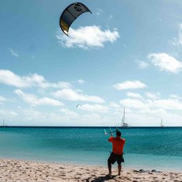 Person kiteboarding on a beach with sailboats in the sea.