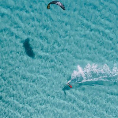 Overhead view of a person kiteboarding on clear turquoise water.