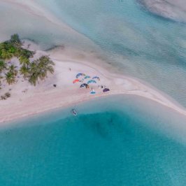 Aerial view of a sandy beach with people under umbrellas near turquoise water and green trees.