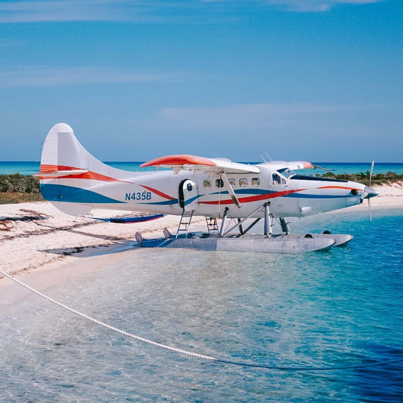 White seaplane with red and blue stripes docked on a sandy beach.
