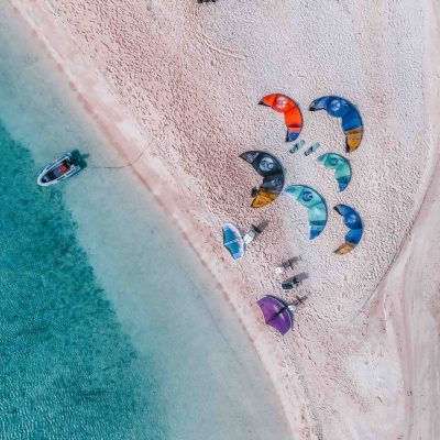 Aerial view of a sandy beach with colorful kites and a boat in turquoise water.