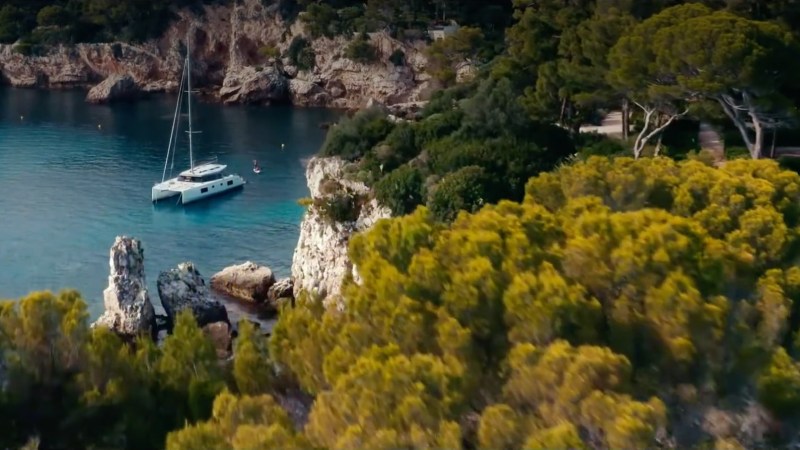 Aerial view of a sailboat on a calm bay surrounded by lush trees and rocky cliffs.