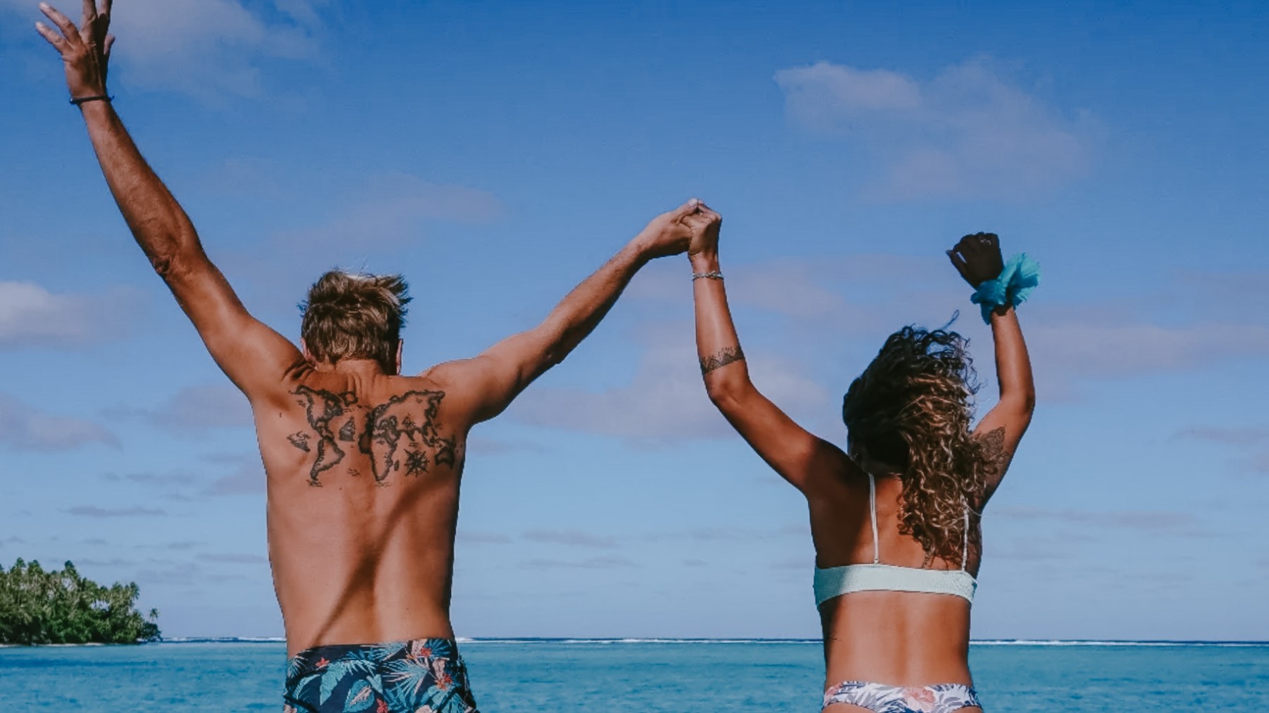 A couple holds hands, facing the ocean, with arms raised on a sunny day.