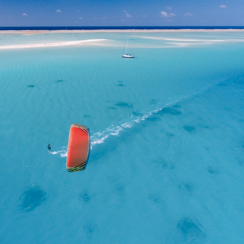 Kitesurfer with red sail rides across turquoise water near a sandy beach with a sailboat.
