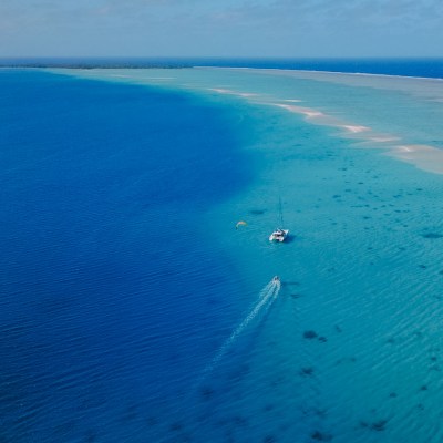 Aerial view of a sailboat and speedboat on clear blue ocean near a sandy shoreline.