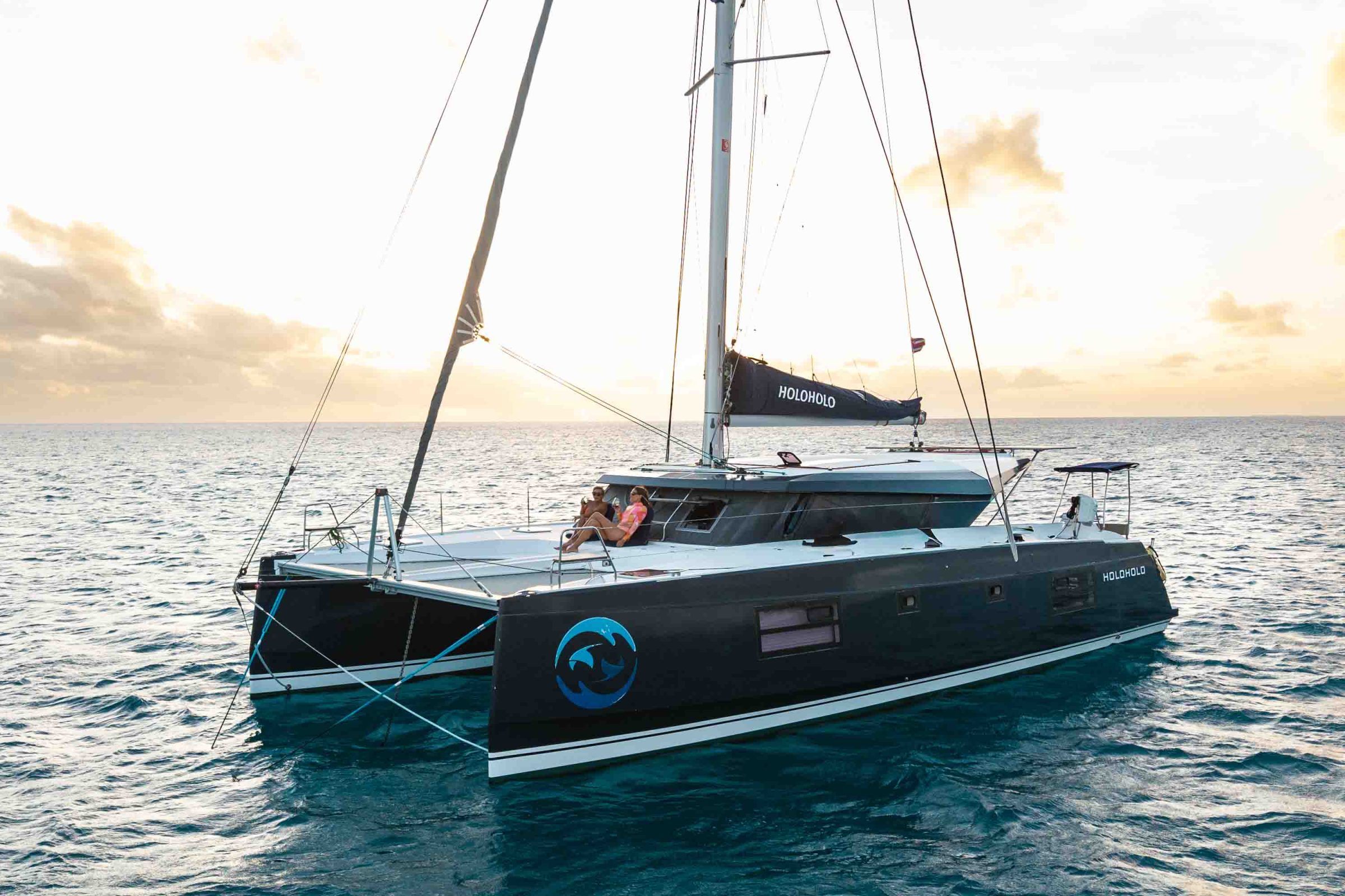 Sailing catamaran on the sea with people relaxing on deck at sunset.