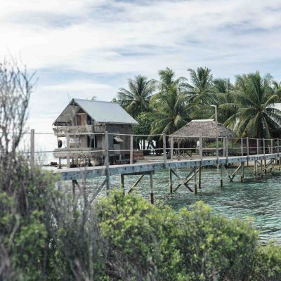 Wooden huts on stilts over clear water with palm trees and cloudy sky.