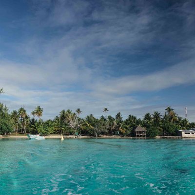 Scenic tropical beach with clear water, palm trees, and buildings under a cloudy sky.
