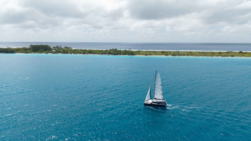 Sailboat on blue ocean near a tree-lined shore under cloudy skies.