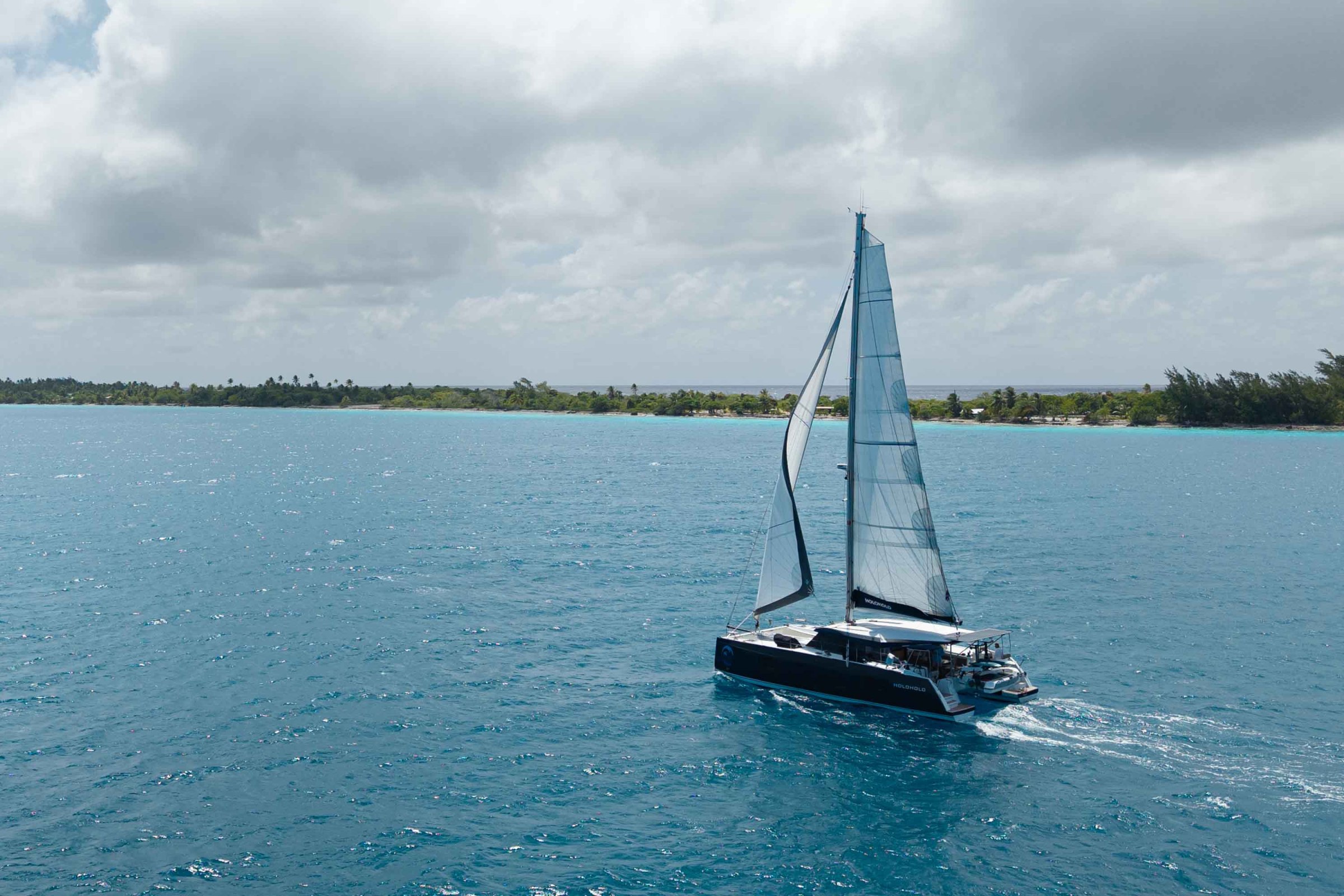 Sailboat on blue ocean near a green shoreline under cloudy skies.