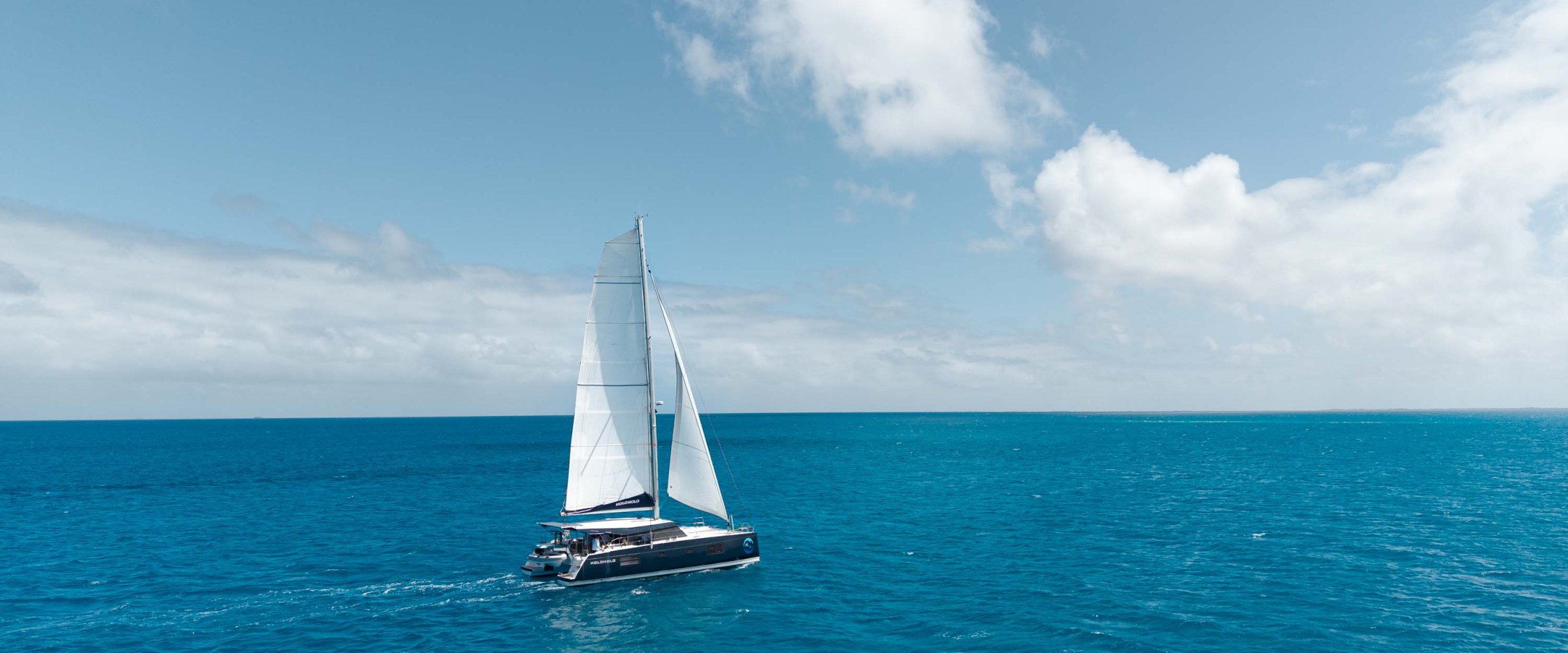 Catamaran sailing on clear blue ocean under sunny skies with scattered clouds.