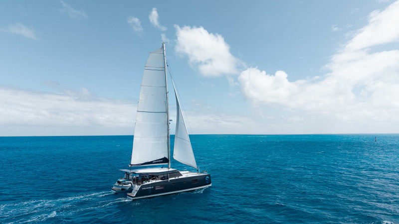 Sailboat with large white sails on a clear blue ocean under a partly cloudy sky.