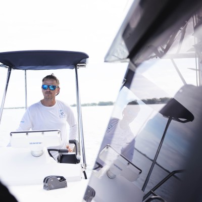 Man with sunglasses steering a boat on a lake, reflection visible on the boat's window.