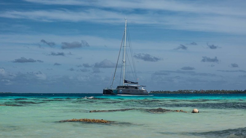 Sailboat anchored near rocky beach with blue sky and clouds.