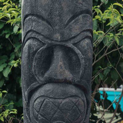 Stone totem pole with carved face and geometric patterns surrounded by foliage.