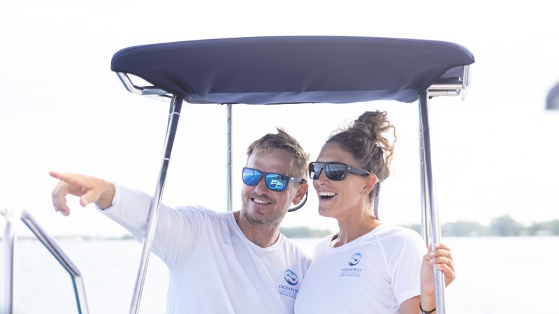 Two people in sunglasses on a boat, one is pointing ahead, both wearing white t-shirts.