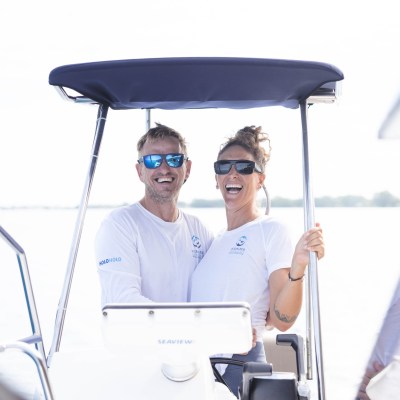 Two people smiling on a boat under a canopy, both wearing sunglasses and white shirts.