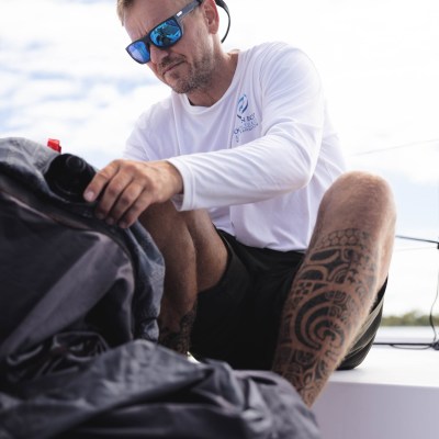 Man in sunglasses and white shirt sitting on a boat with a large backpack.