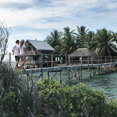 Two people walking on a wooden pier over clear water towards huts and palm trees.