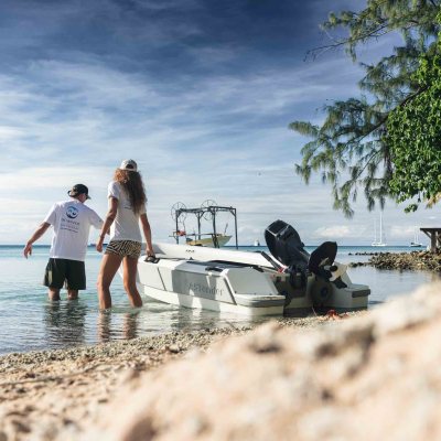 Two people walk a boat in shallow beach water, surrounded by trees and a bright sky.
