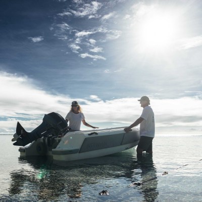 Two people stand by a small boat in shallow water under a bright sun.